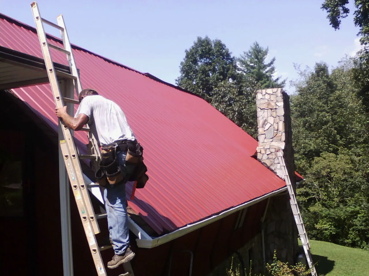 Roofer on a ladder beside a red metal roof and stone chimney — Soffit & Fascia Repair by Roofing Brothers in Scotia